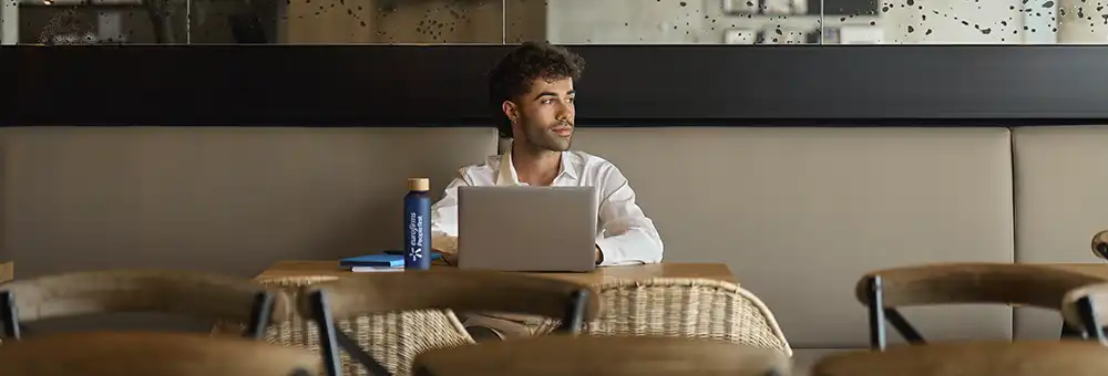 Pessoa a trabalhar num portátil numa mesa de café, com garrafa reutilizável e caderno ao lado.